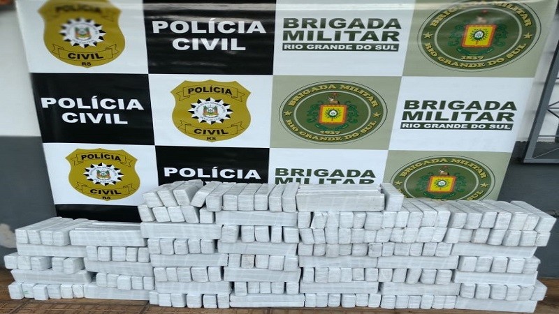 Mais de 200 Kg de maconha empilhados e expostos ao chão em frente ao banner da Brigada Militar e da Polícia Civil