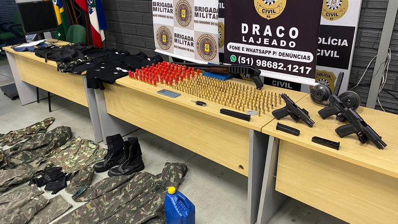 Foto mostra três mesas com munições e armas de fogo. No chão, na frente da mesa, há uniformes militares.