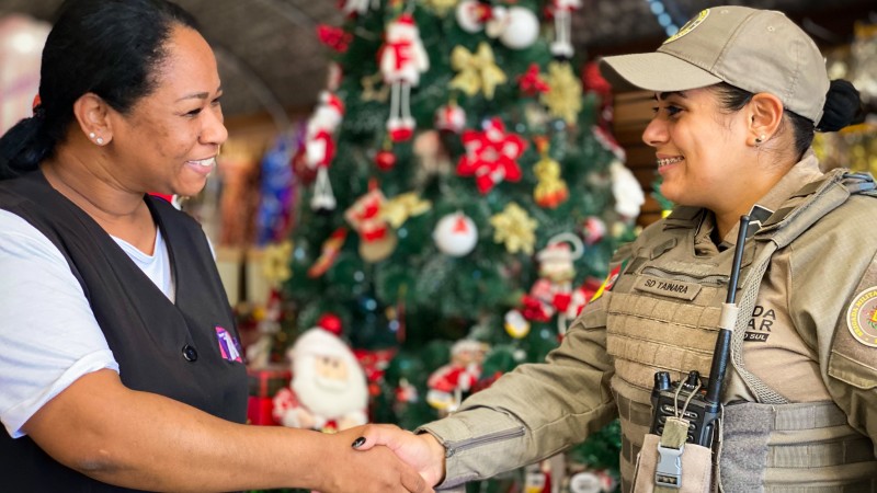 Mulher cumprimenta policial militar feminina em estabelecimento comercial. 