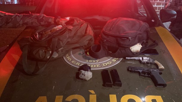 Imagem mostra duas mochilas e duas pistolas em cima do capô de uma viatura da BM.