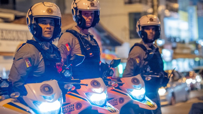 Foto mostra três policiais militares da Rocam em pé, à noite, com suas motos com farol ligado ao lado deles
