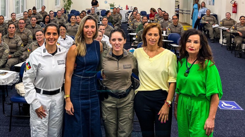 A imagem mostra as palestrantes do evento em pé, com as brigadianas atrás delas posando para foto