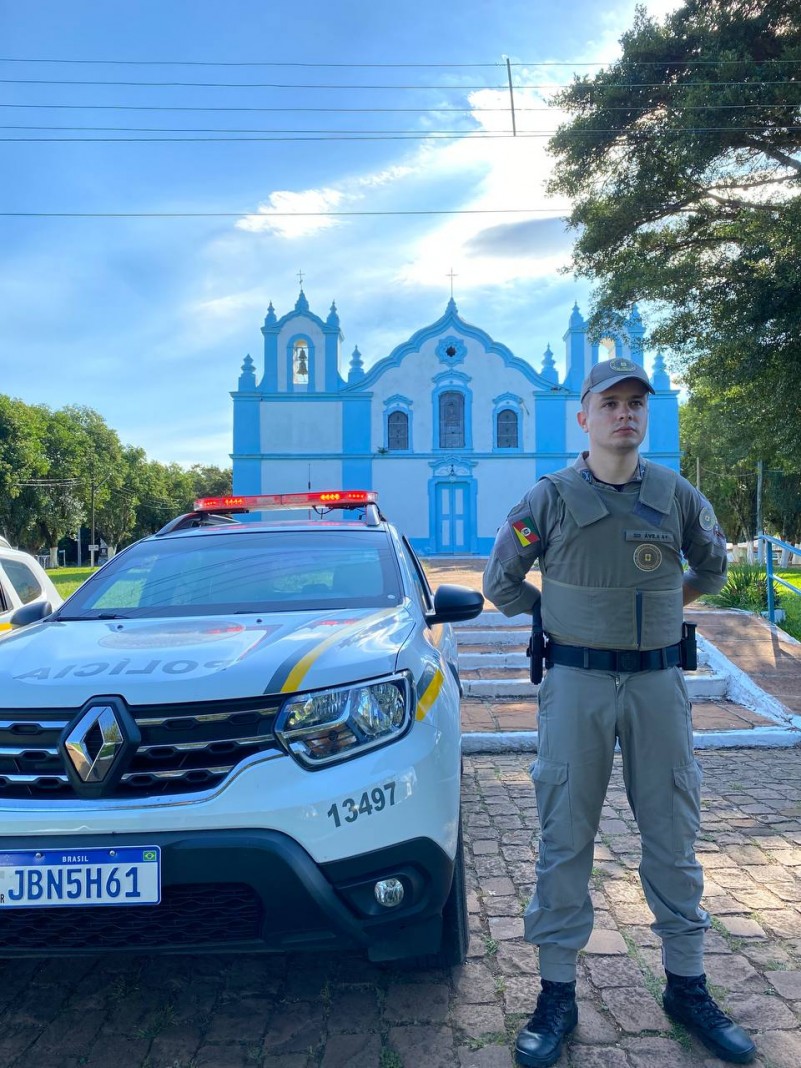 Um policial militar ao lado de uma viatura, em frente a uma igreja