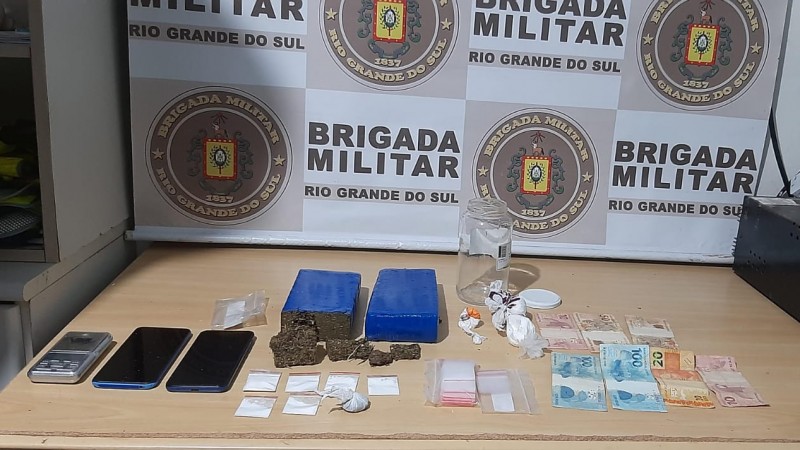 Ao fundo, banner da Brigada Militar e sobre a mesa quase dois quilos de maconha e porções de cocaína apreendidas