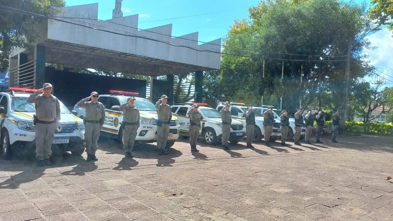 13 policiais militares em frente a viaturas, prestando continência ao terreno.