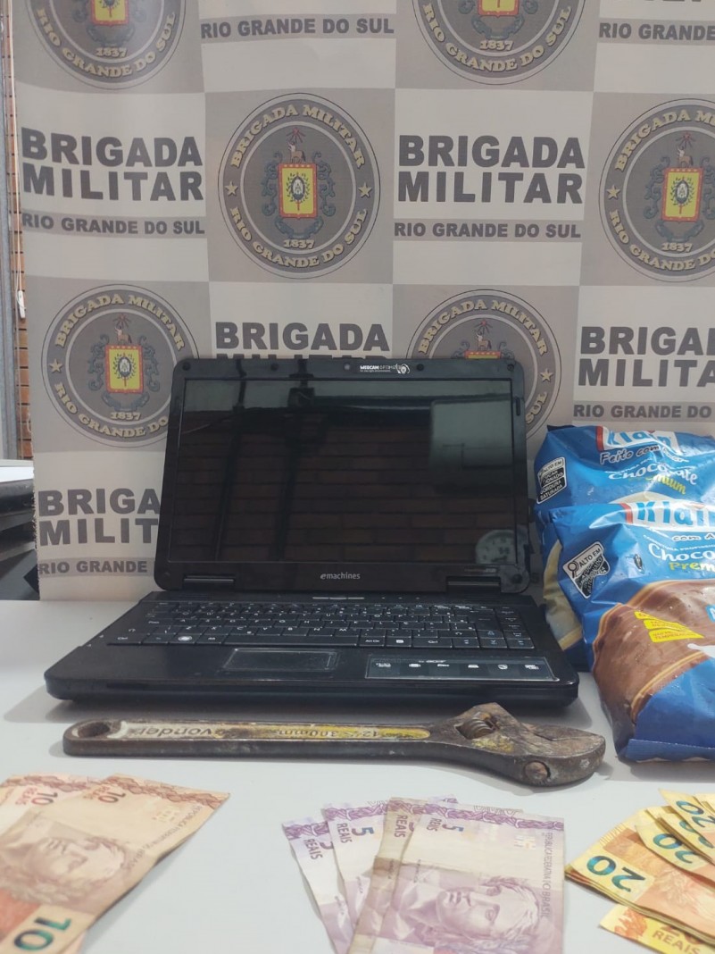 Sobre uma mesa de cor cinza, estão os materiais recuperados, um notebook, um valor em dinheiro e dois pacotes de chocolate. Ao fundo está o banner da Brigada Militar.