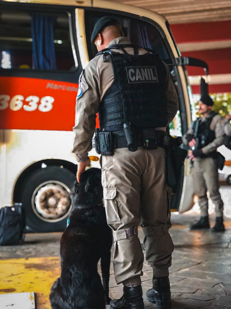 Policial do canil do CRPO Litoral, ao lado de seu cão de faro. Ao fundo, ônibus na rodoviária