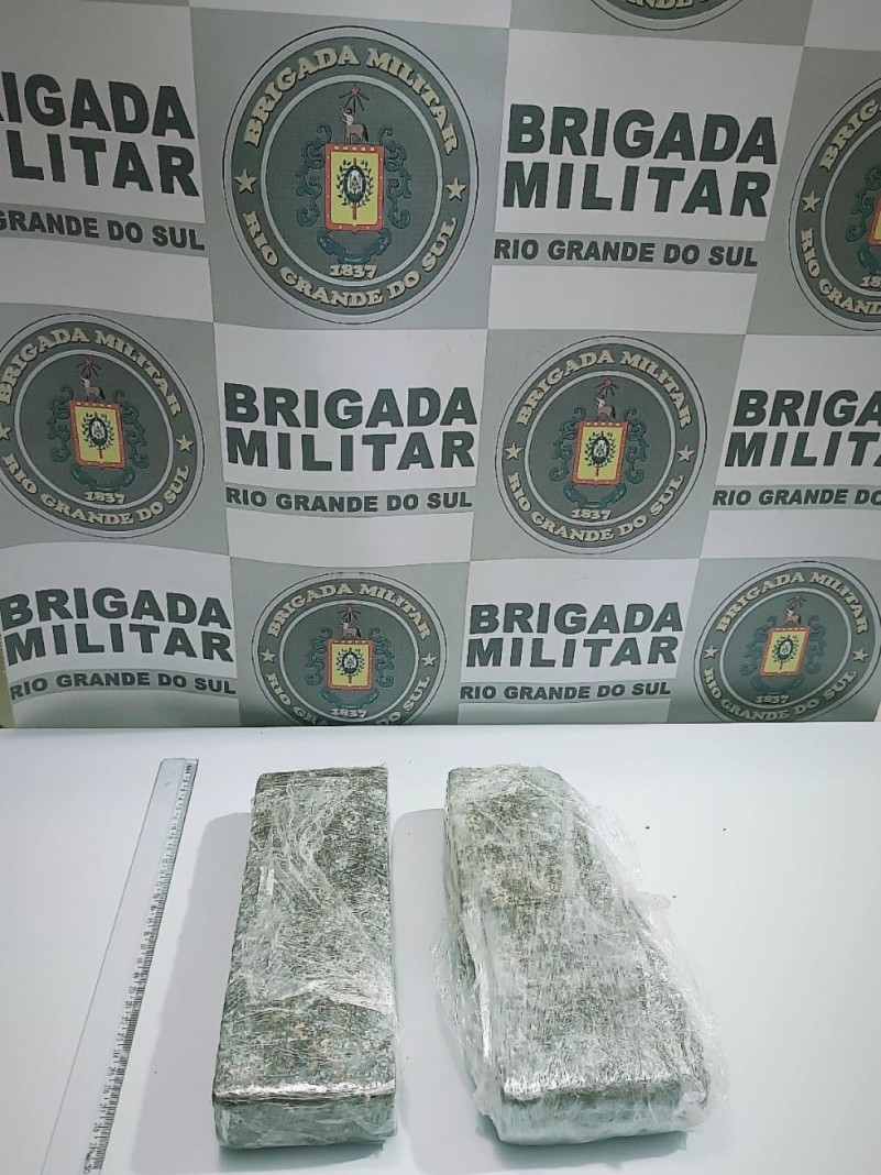 Na foto, sobre uma mesa de cor cinza, est&atilde;o dois "tijolos" de maconha, que pesam 1,565 Kg, subst&acirc;ncia psicoativa, de cor esverdeada, que encontra-se embalada em papel filme transparente. Ao fundo est&aacute; o banner da Brigada Militar.