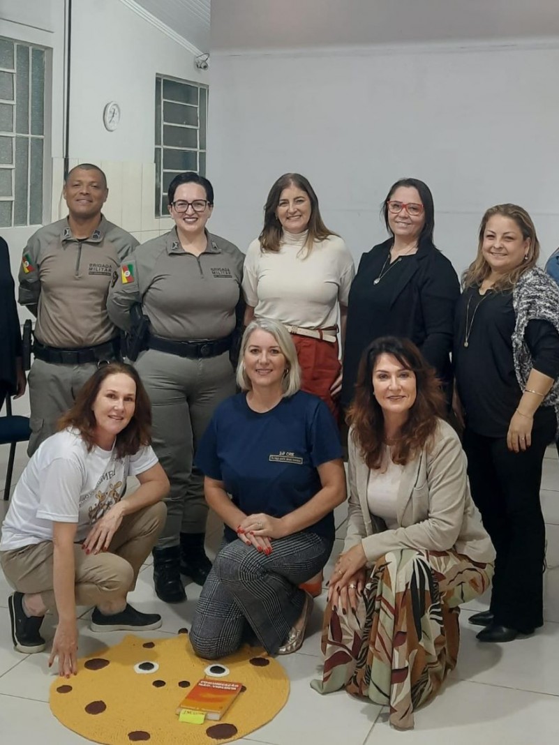 Na foto ao fundo temos uma parede de cor branca, com duas janelas de vidro do  lado esquerdo da sala. Mais a frente, postadas em p&eacute;, encontram-se algumas pessoas, dentre elas dois policiais militares. Na ocasi&atilde;o ocorreu uma reuni&atilde;o integrada com os membros do N&uacute;cleo Municipal de Pr&aacute;ticas Restaurativas (NUPRA), em prol de uma cultura de paz nas escolas.