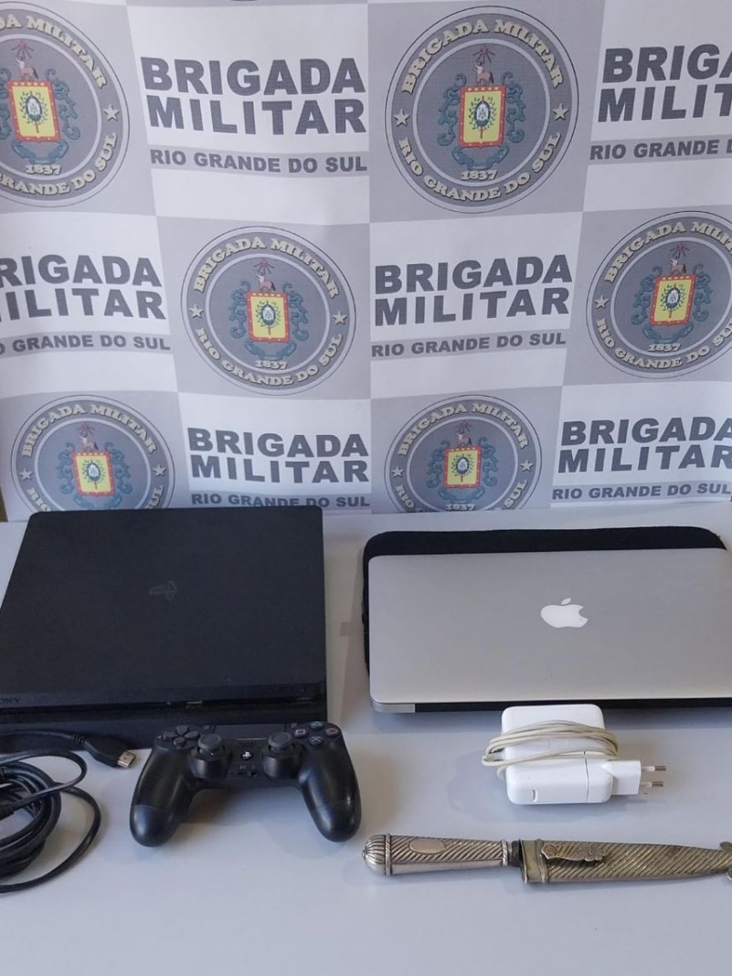 Na foto, em cima de uma mesa de cor branca, encontram-se um 01 MacBook marca APPLE de cor branca, 01 V&iacute;deo game PLAYSTATION 4 de cor preta e uma faca de prata, todos materiais de furto que foram recuperados. Ao fundo, temos o banner da Brigada Militar.