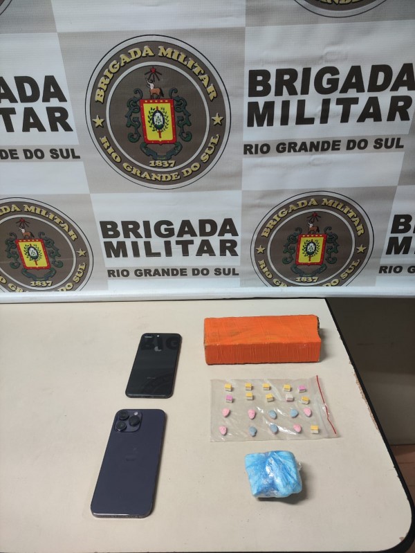 um tijolo de maconha e 20 comprimidos de ecstasy em cima de uma mesa, ao fundo banner da Brigada Militar