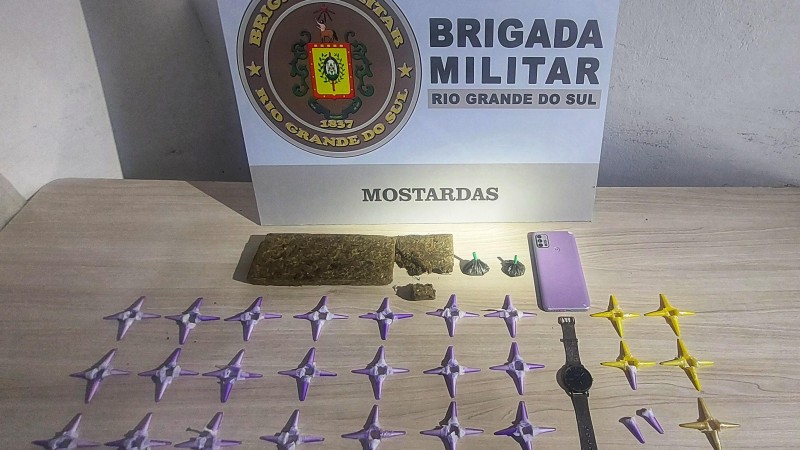 Em cima de uma mesa, 102 pinos de cocaína e um tijolo de maconha. Ao fundo um banner da Brigada Militar