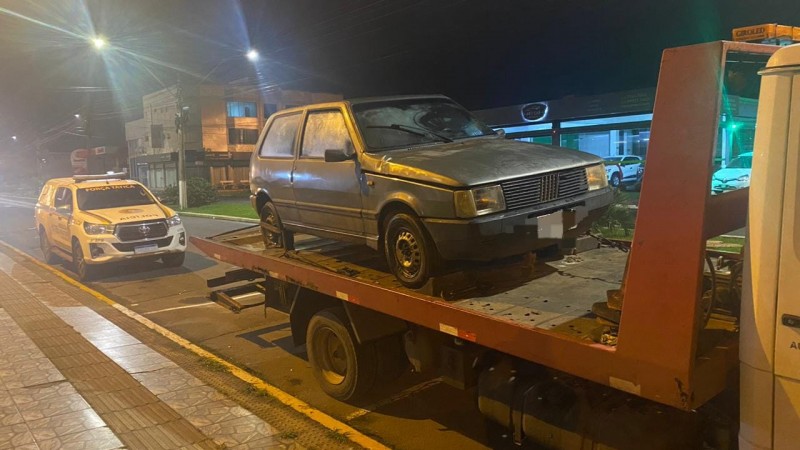 Na imagem há um veículo Fiat Uno cor cinza, em cima de um guincho. Ao fundo, uma viatura da Força Tática do 8º BPM