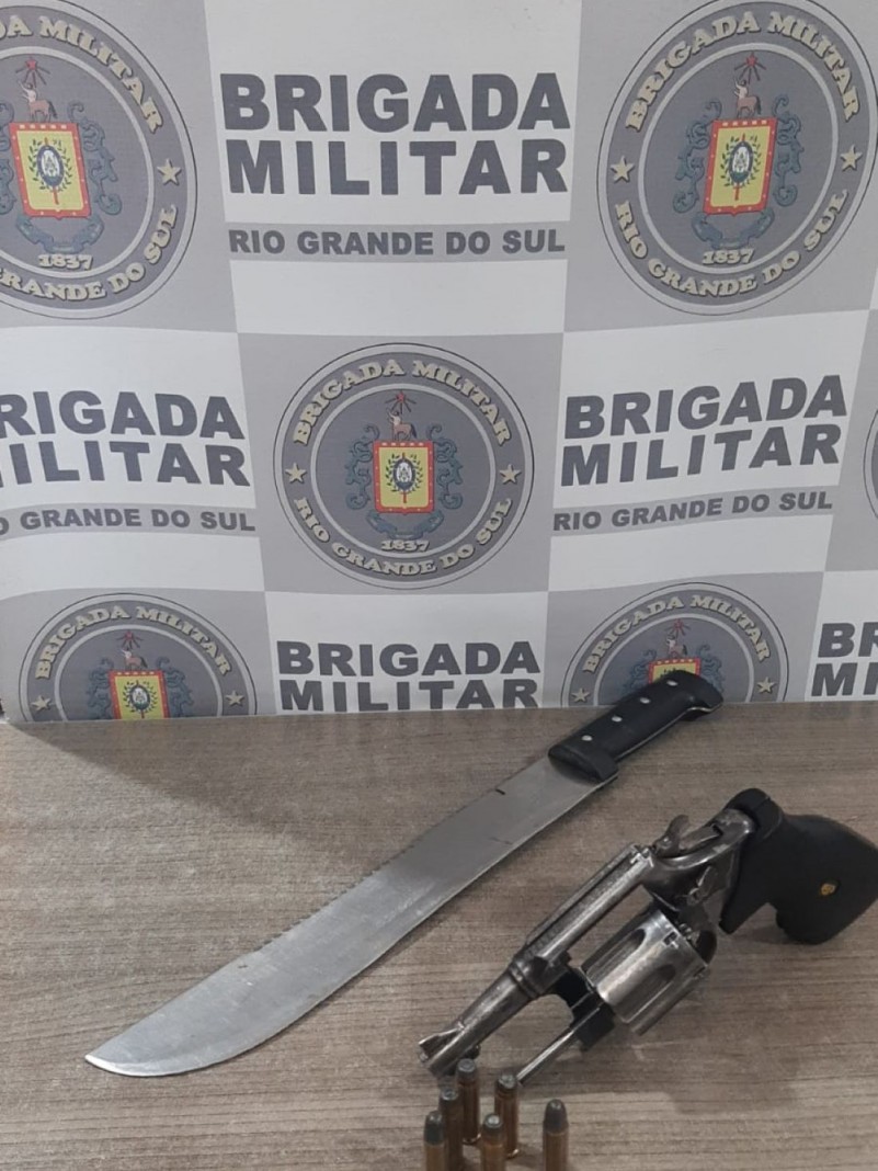 Na foto sobre uma mesa de cor marrom, est&aacute; posicionada uma arma de fogo, modelo rev&oacute;lver de calibre 38, com seis muni&ccedil;&otilde;es de mesmo calibre, e um fac&atilde;o com l&acirc;mina de alum&iacute;nio de 25 cent&iacute;metros e cabo de cor preta. Ao fundo encontra-se o banner da Brigada Militar.