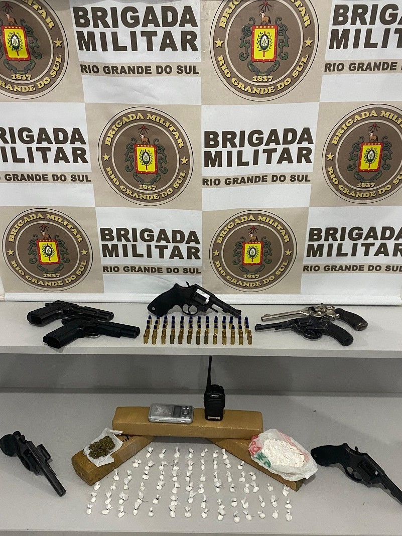 Sete armas, drogas e munições sobre uma mesa com banner da BM ao fundo
