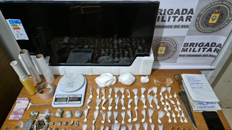 Ao fundo, banner institucional da Brigada Militar, televisão apreendida, caderno de anotações do tráfico, 68 porções de cocaína, 22 porções de maconha, duas pedras de crack, cinco munições de calibre 38, balança de precisão e material de embalar drogas