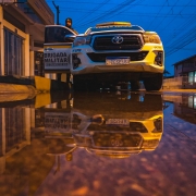 Na imagem há um policial militar da Força Tática do 8ºBPM ao lado de uma viatura. Há o reflexo da imagem em uma poça de água