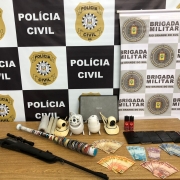 Ao fundo banner das instituições (Brigada Militar e Polícia Civil) e em cima de uma mesa as apreensões - espingarda de pressão, quatro câmeras de monitoramento, seis celulares, um notebook, materiais para embalagem, spray de pimenta, uma balança de precisão e um valor de R$ 1390.