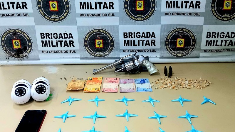 Em cima de uma mesa, um revólver calibre 38, cinco munições, 88 pedras de crack, 63 pinos de cocaína, duas câmeras de segurança e ao fundo o banner da Brigada Militar