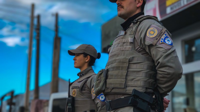 Dois policiais (um masculino em primeiro plano e uma feminina em segundo plano) em frente a um estabelecimento