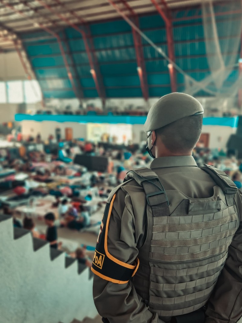 Foto mostra policial militar em pé em primeiro plano. Em segundo plano, um abrigo durante enchente no Rio Grande do Sul.