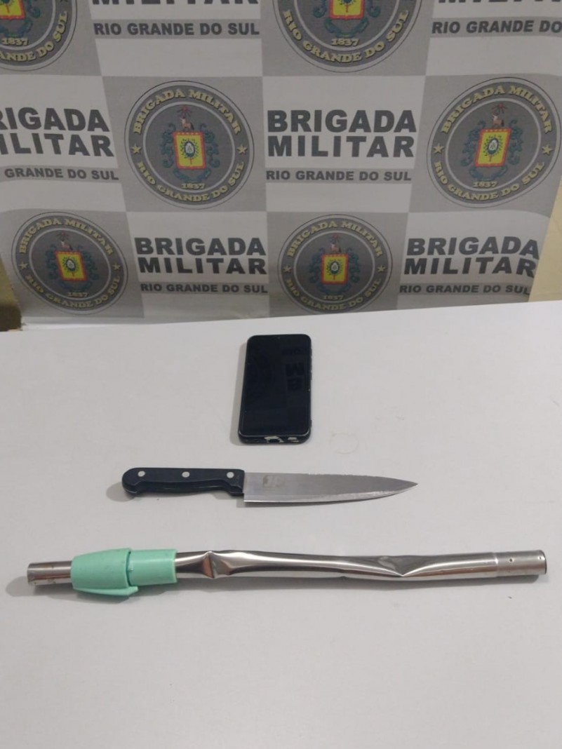 Na foto, ao fundo temos o banner Institucional da Brigada Militar, e à frente sobre uma mesa de cor branca, está um celular de cor preta, uma faca com cabo preto com lâmina em alumínio, e uma barra de ferro.