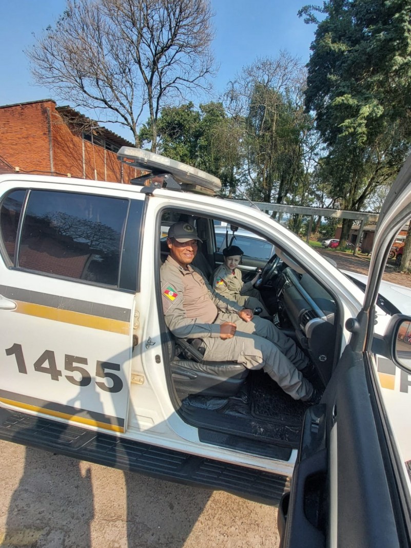 Na foto ao fundo temos o céu azul e algumas árvores do centro para o lado direito. Mais à frente está uma viatura da Brigada Militar, em cor Branca com adesivos de cor amarelo e cinza. Dentro dela encontram-se um policial militar e o pequeno Cassiano, um menino de 5 anos que é admirador da Brigada Militar.