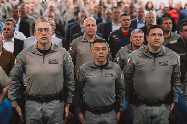 Foto mostra os três oficiais do Comando da BM perfilados durante execução do Hino Nacional Brasileiro, dentro de um auditório