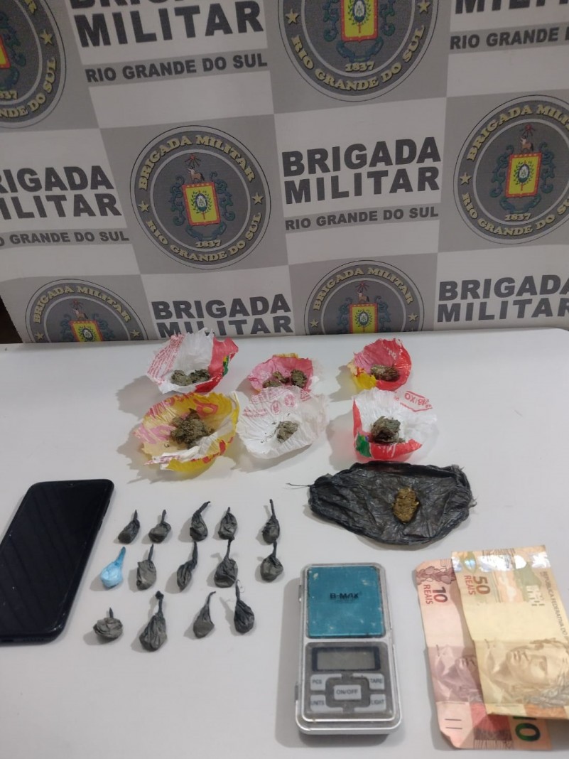 Ao fundo temos o Banner Institucional da Brigada Militar. Mais à frente sobre uma mesa de cor branca, estão 07 porções de maconha( substância química psicoativa, de cor esverdeada), 14 porções de cocaína (substância química psicoativa, de cor branca), sessenta reais em espécie, uma balança de precisão, e um telefone celular de cor preta. Todos os materiais foram apreendidos em Cachoeira do Sul.