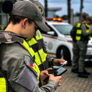 Na imagem, dois alunos-soldados mais a frente, verificando documentos no sistema operacional de consultas e, mais ao fundo, dois militares e uma viatura, em policiamento ostensivo, fazendo a segurança dos outros alunos-soldados. 