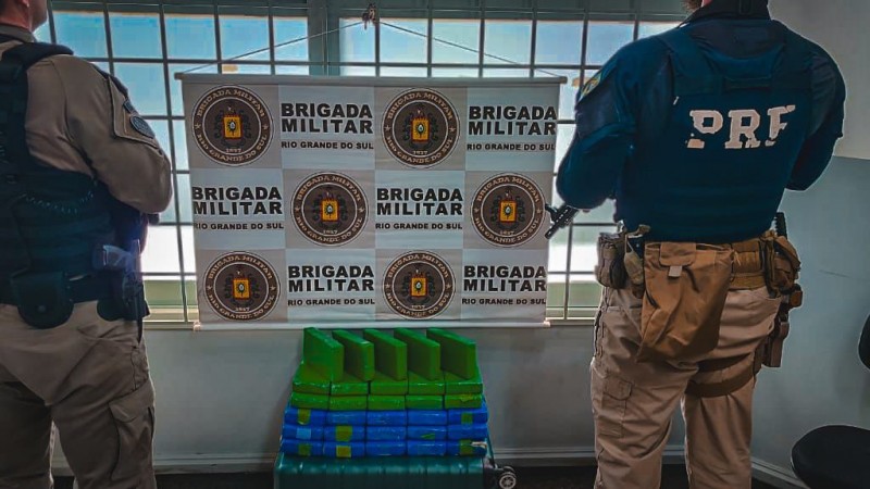 Na imagem, um militar da BM à esquerda, um policial da PRF à direita, e, ao centro, o banner da BM e as drogas apreendidas a frente