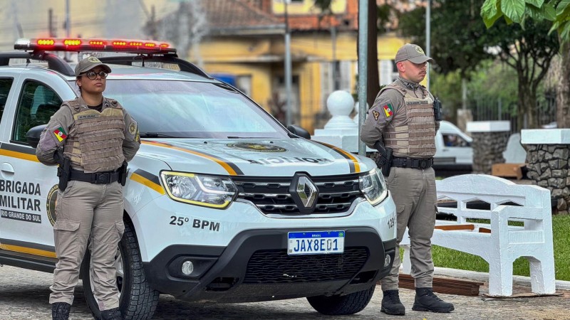 Uma dupla de policiais militares, sendo um homem e uma mulher, a frente de uma viatura da Brigada MIlitar em um praça em São Leopoldo