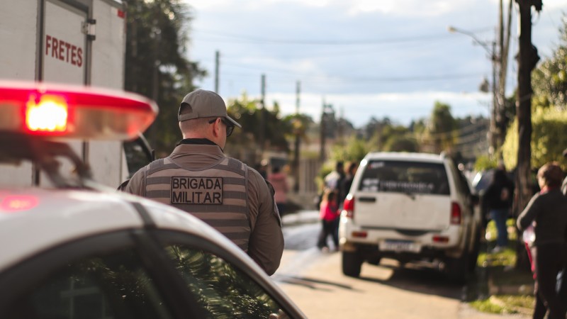 Foto mostra uma viatura parada com o giroflex ligado em uma avenida. Mais à frente, movimentação de pessoas e um carro branco parado.