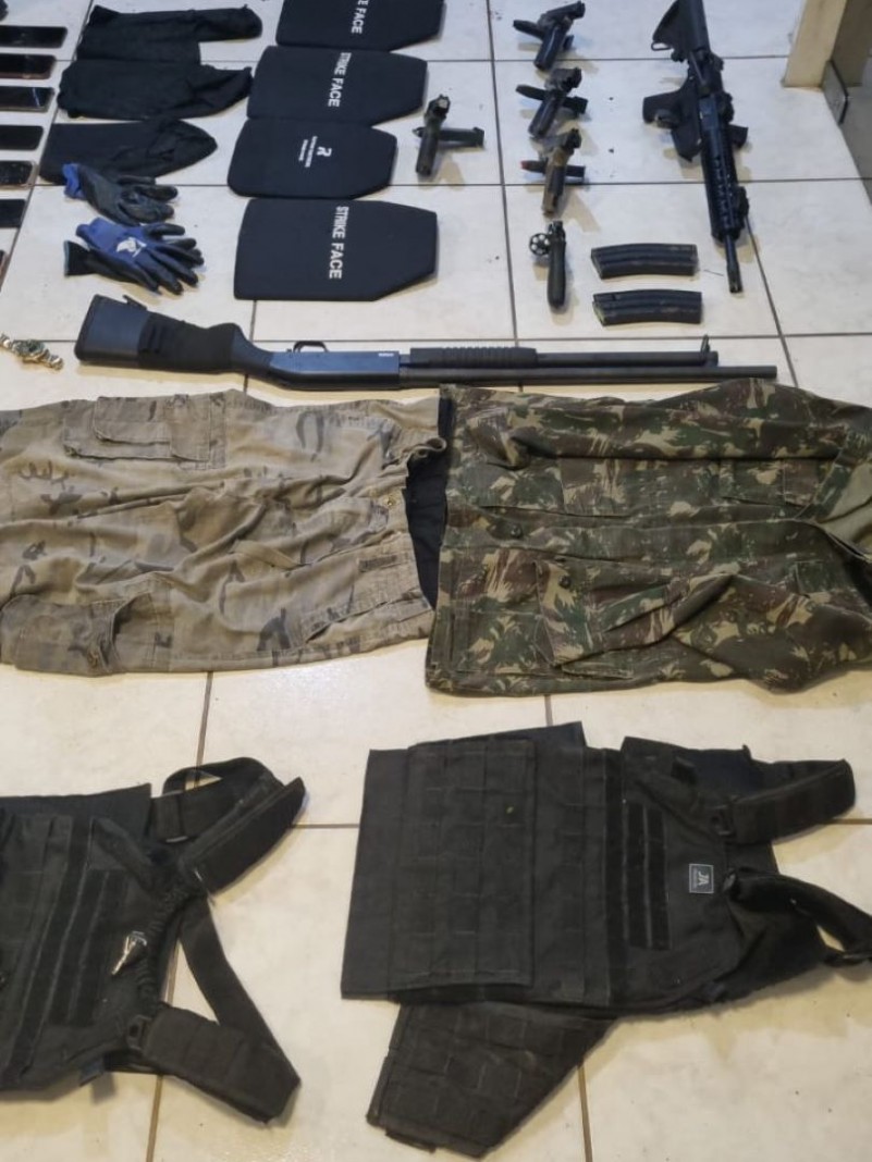 Material apreendido é espalhado no chão, estão um fuzil, quatro pistolas, um revolver, roupas, placas balisticas, celulares e uma escopeta.