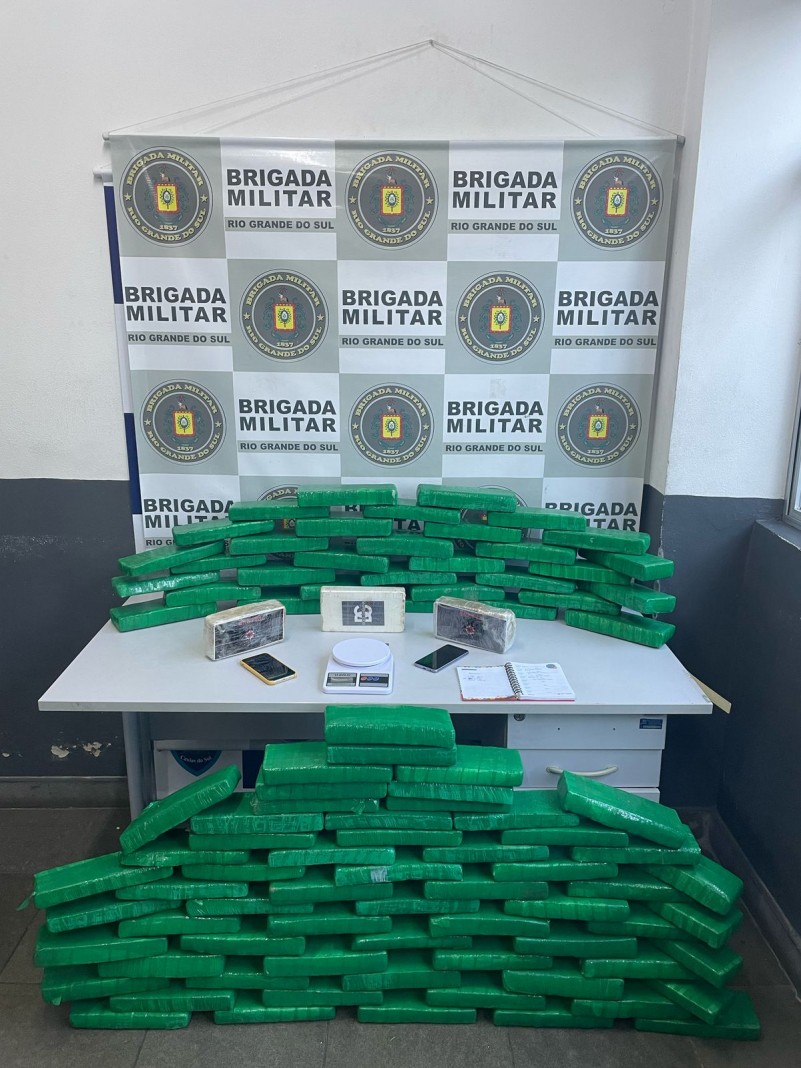 Foto mostra quase uma centena de porções de drogas empilhadas em cima e embaixo de uma mesa, com banner da Brigada Militar ao fundo