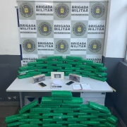 Foto mostra quase uma centena de porções de drogas empilhadas em cima e embaixo de uma mesa, com banner da Brigada Militar ao fundo