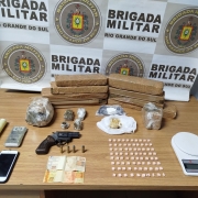 Foto mostra porções de drogas empilhadas em cima de uma mesa, junto a balanças de precisão, celular, notas de dinheiro e uma arma. Em segundo plano, o banner da Brigada Militar.