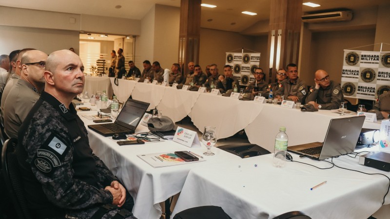 Foto mostra oficiais atentos a uma apresentação, enquanto estão sentados a uma mesa retangular.