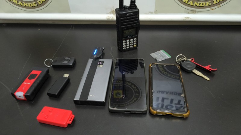 Na foto contém um radio na frequência da BM, 01 aparelho com 02 pen-drives bloqueador de sinal, 01 imã desbloqueio porta de veículos, 02 controles diversos, 01 aparelho p/dar partida em veículo, 02 aparelhos celulares.
