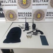 Uma arma de fogo, munições, uma touca, dois radios portáteis e um aparelho de celular em uma mesa em frente ao banner da Brigada Militar.