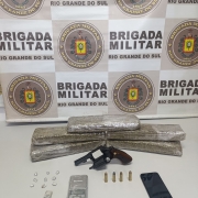 Porções de droga, uma arma de fogo, uma balança pequena, munições e um aparelho celular em frente ao banner da Brigada Militar.