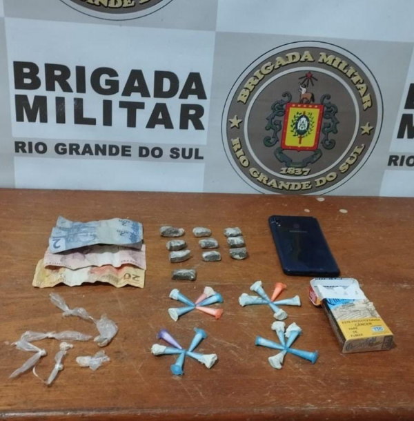 20 pinos de cocaína, sete pedras de crack, 10 porções de maconha dispostos sob uma mesa e ao fundo banner da Brigada Militar