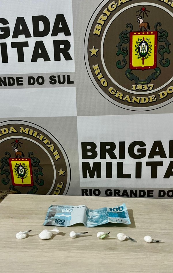 Seis porções de cocaína e 100 reais e ao fundo banner da Brigada Militar