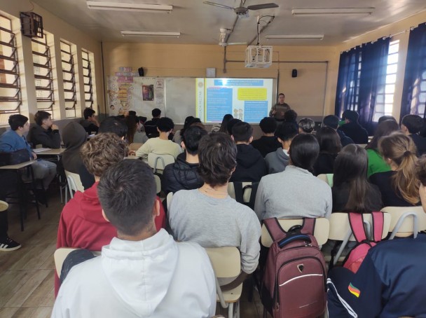 Sala de aula lotada de estudantes. Ao fundo, uma policial militar ministra uma palestra e aparecem slides expostos no quadro. 