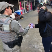 Policial militar feminina conversa com uma mulher caminhando na rua e entrega um folder da Patrulha Maria da Penha 