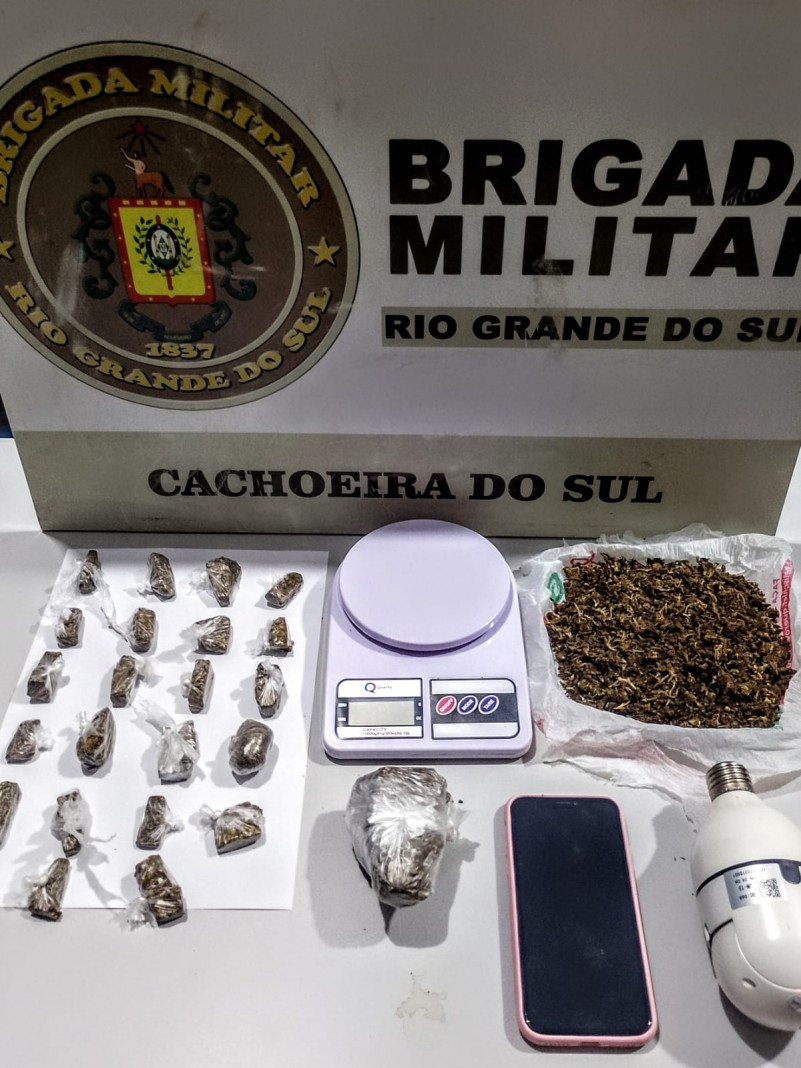 Na imagem sobre uma mesa 01 por&ccedil;&atilde;o grande de maconha; 22 por&ccedil;&otilde;es pequenas fracionadas de maconha; 01 por&ccedil;&atilde;o de maconha tipo Skank; 01 aparelho celular; 01 c&acirc;mera; e 01 balan&ccedil;a de precis&atilde;o.