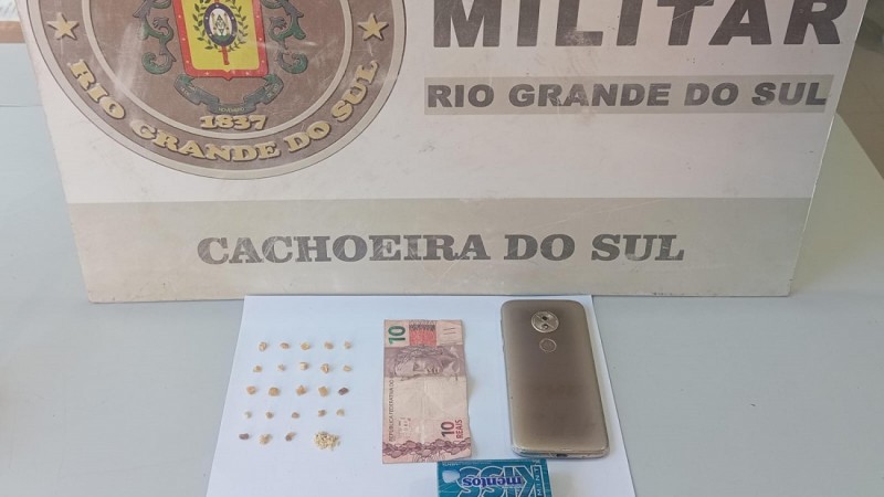 Na imagem sobre uma mesa 24 por&ccedil;&otilde;es de crack pesando 2g;  01 celular marca Motorola na cor dourado; 01 pote azul marca Mentos e R$ 10,00 em moeda corrente, ao fundo o banner institucional da BM.