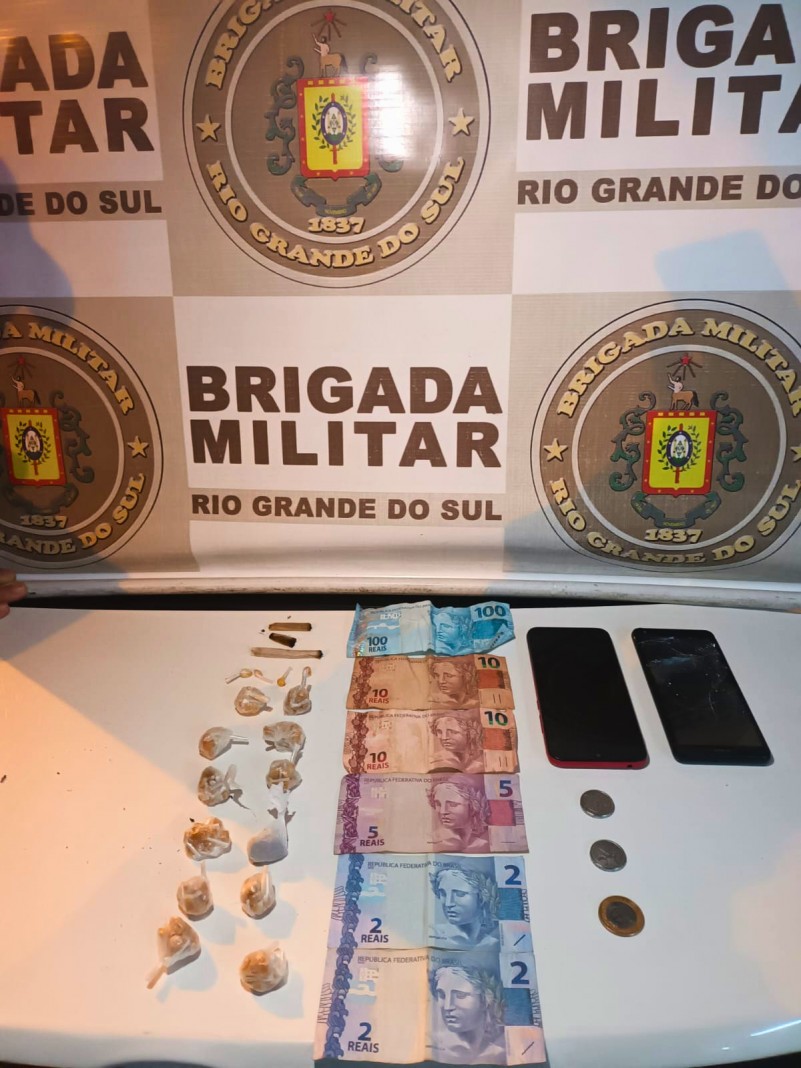 Banner Brigada Militar sobre mesa com exposi&ccedil;&atilde;o dos materiais apreendidos: 150 por&ccedil;&otilde;es de crack, quatro por&ccedil;&otilde;es de maconha, dois telefones celular e valor em esp&eacute;cie.