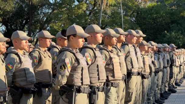 Imagem mostra policiais militares alinhados durante formatura ao ar livre. Os integrantes utilizam uniforme operacional, colete balístico e boné institucional, com árvores ao fundo.