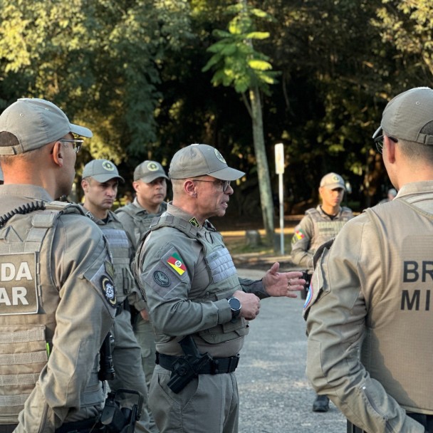Imagem mostra o subcomandante-geral da Brigada Militar, coronel Jorge Dirceu Abreu Silva Filho, conversando com policiais militares durante a formatura de lançamento da Operação Convergência. Os integrantes utilizam uniforme operacional cáqui, colete balístico e boné institucional. Ao fundo, aparecem árvores do Parque Farroupilha, em Porto Alegre.